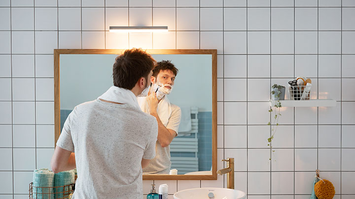 Man shaving in front of a bathroom mirror lighted by a wall mounted Philips LED